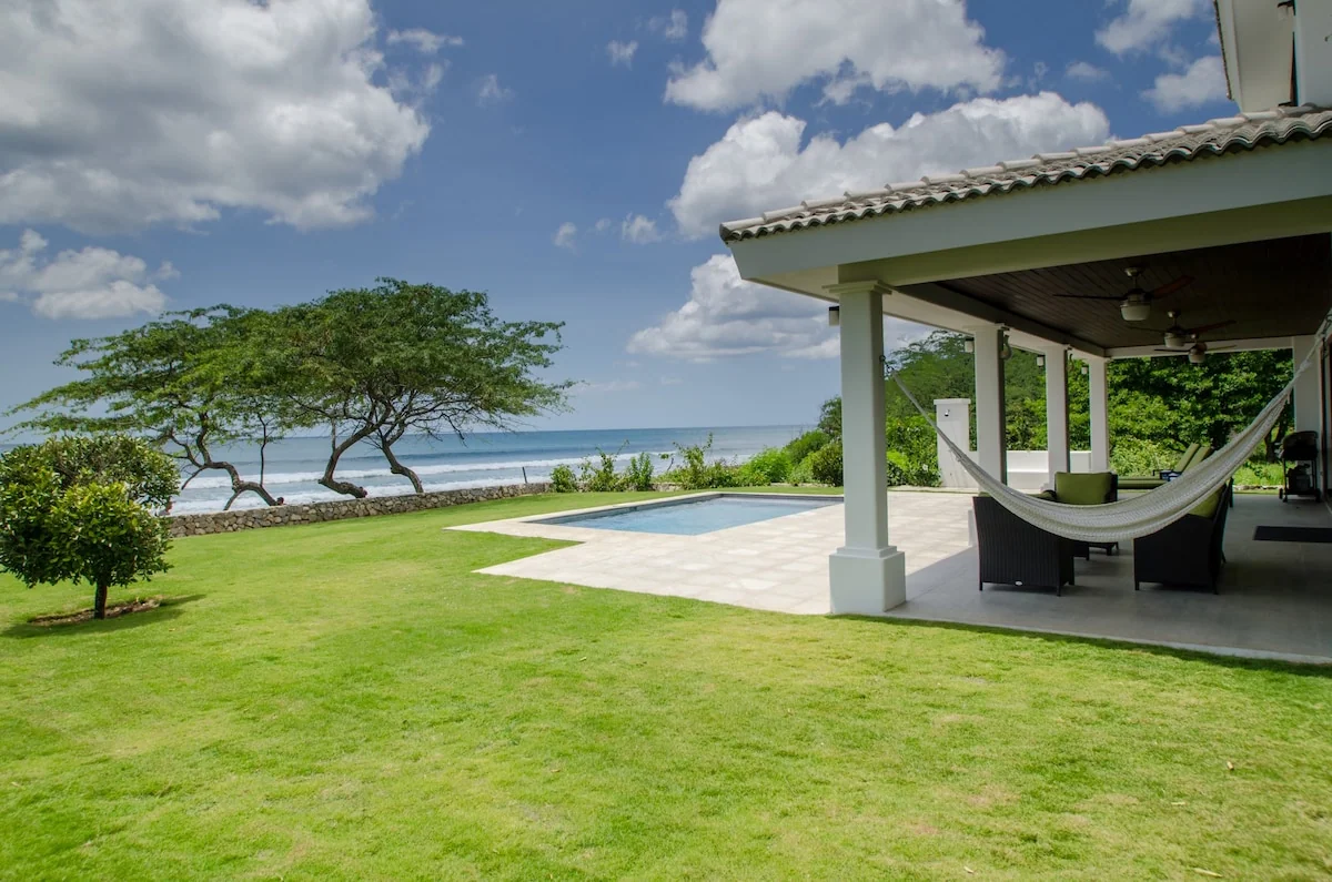 A sunny coastal property shows a covered patio with a hammock, a lawn, a pool, trees, and the ocean beyond.
