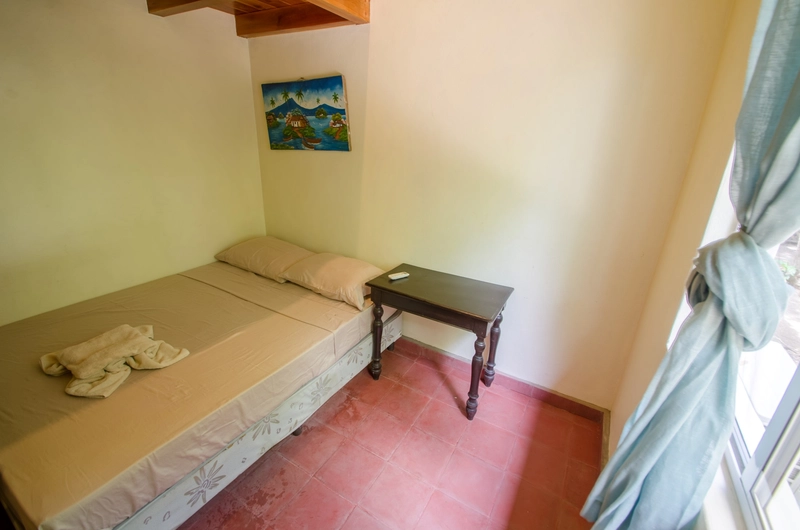 The image depicts a small bedroom with a single bed positioned against a light-colored wall. The bed is dressed with beige linens and two pillows. A folded towel is placed on the b