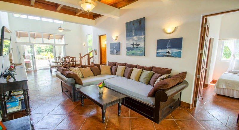 The image shows a spacious and bright living room with a large L-shaped sofa adorned with multiple cushions. A small wooden coffee table with a vase of flowers is in front of the s