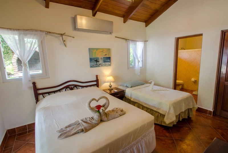 The image shows a cozy bedroom with two beds. The larger bed features neatly folded towels in the shape of swans forming a heart, while the smaller bed also has a towel folded into