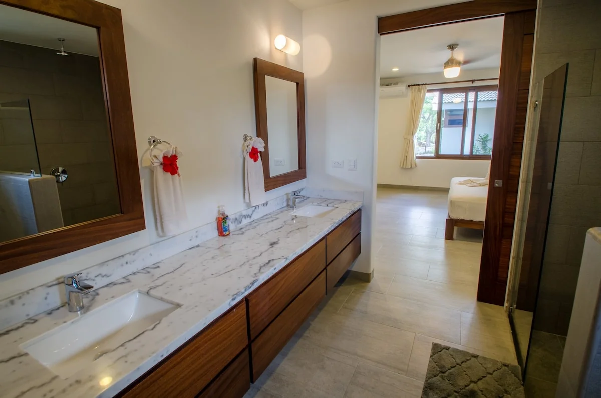 A spacious bathroom with a marble vanity, large mirrors, wall sconces, towels, and an open doorway leading to a bedroom.