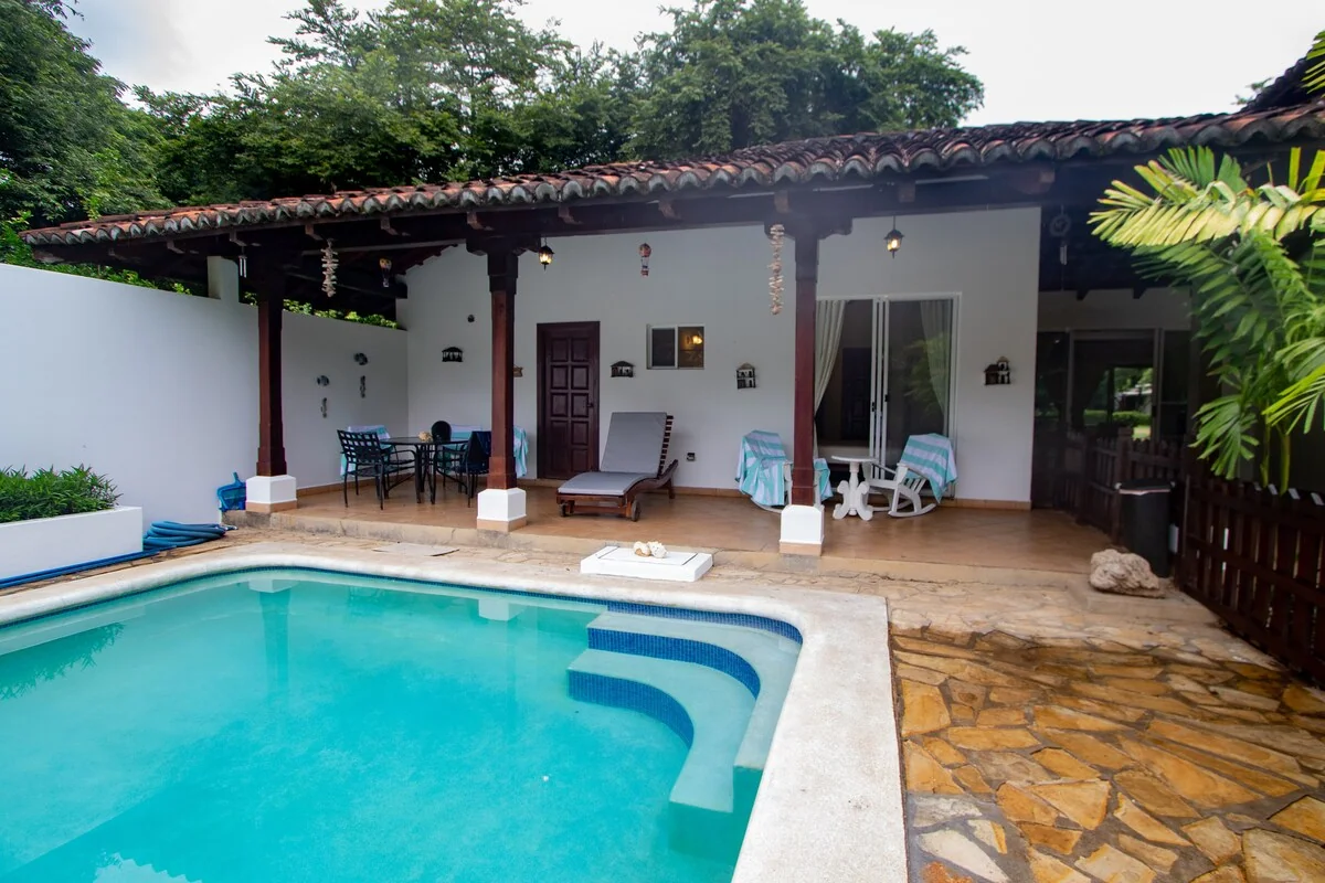 A turquoise swimming pool sits beside a covered patio with seating, loungers, and a white-walled house.
