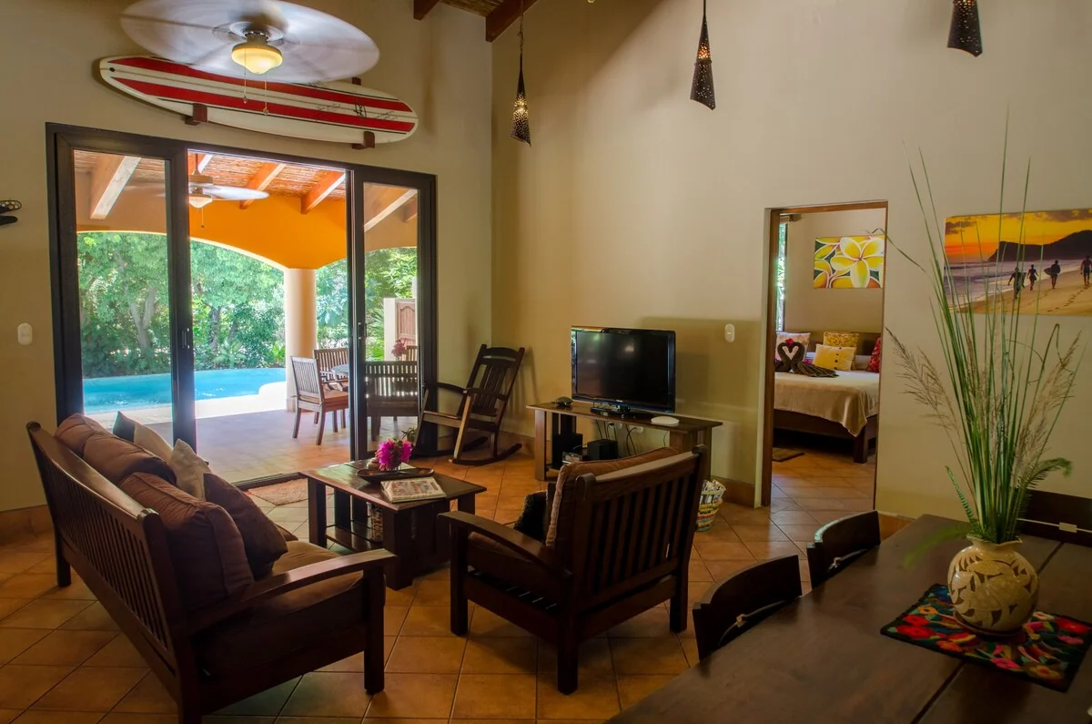 A warmly furnished living room opens onto a covered patio and swimming pool, with a television, seating, plants, and a bedroom visible through a doorway.
