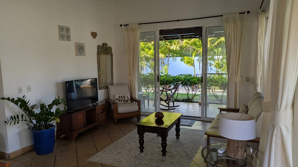 A bright living room with a television, coffee table, armchairs, plants, and glass doors opening toward greenery outside.