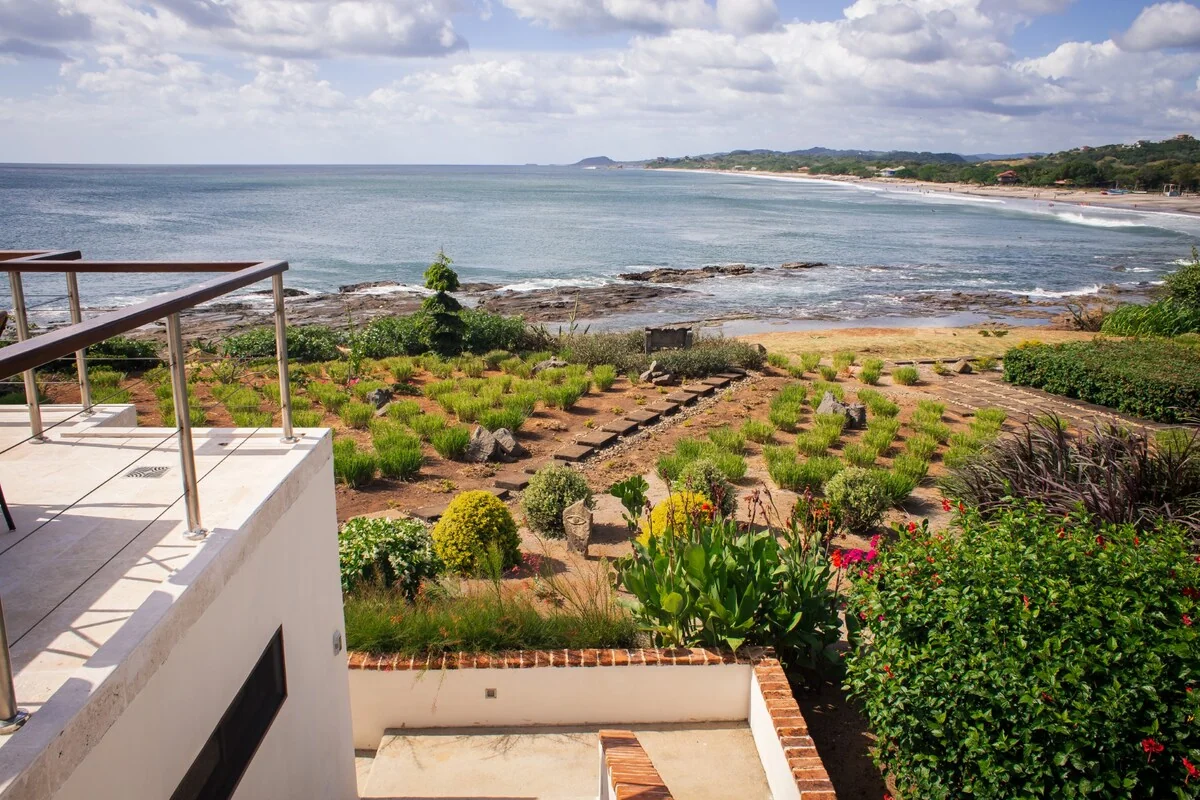 A landscaped garden and terrace overlook a rocky shoreline, calm ocean, and distant tree-lined coast beneath a partly cloudy sky.