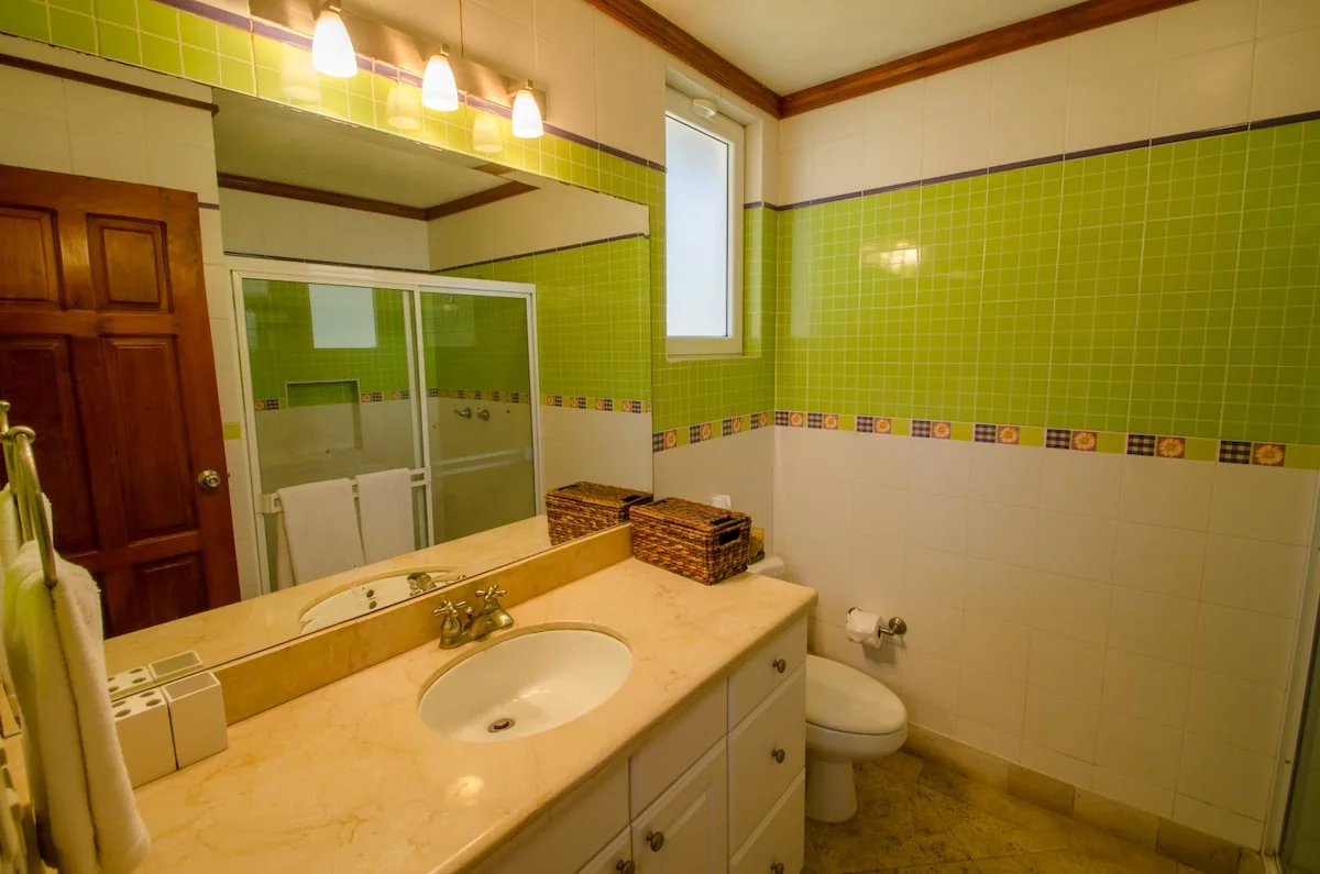 A brightly lit bathroom with green tiled walls, a large vanity mirror, sink, toilet, and glass-enclosed shower.