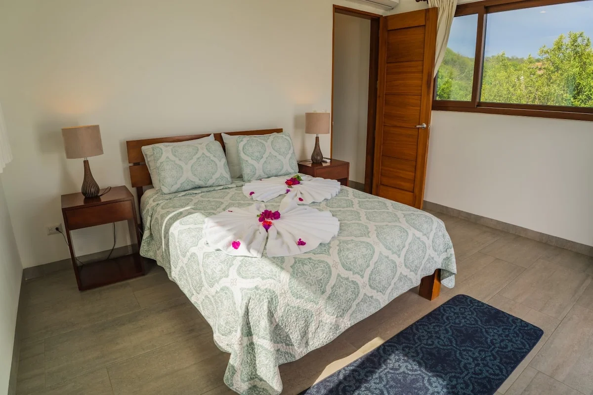 A neatly made bedroom features a patterned bedspread with flower petals, bedside lamps, a wooden door, and a window overlooking greenery.