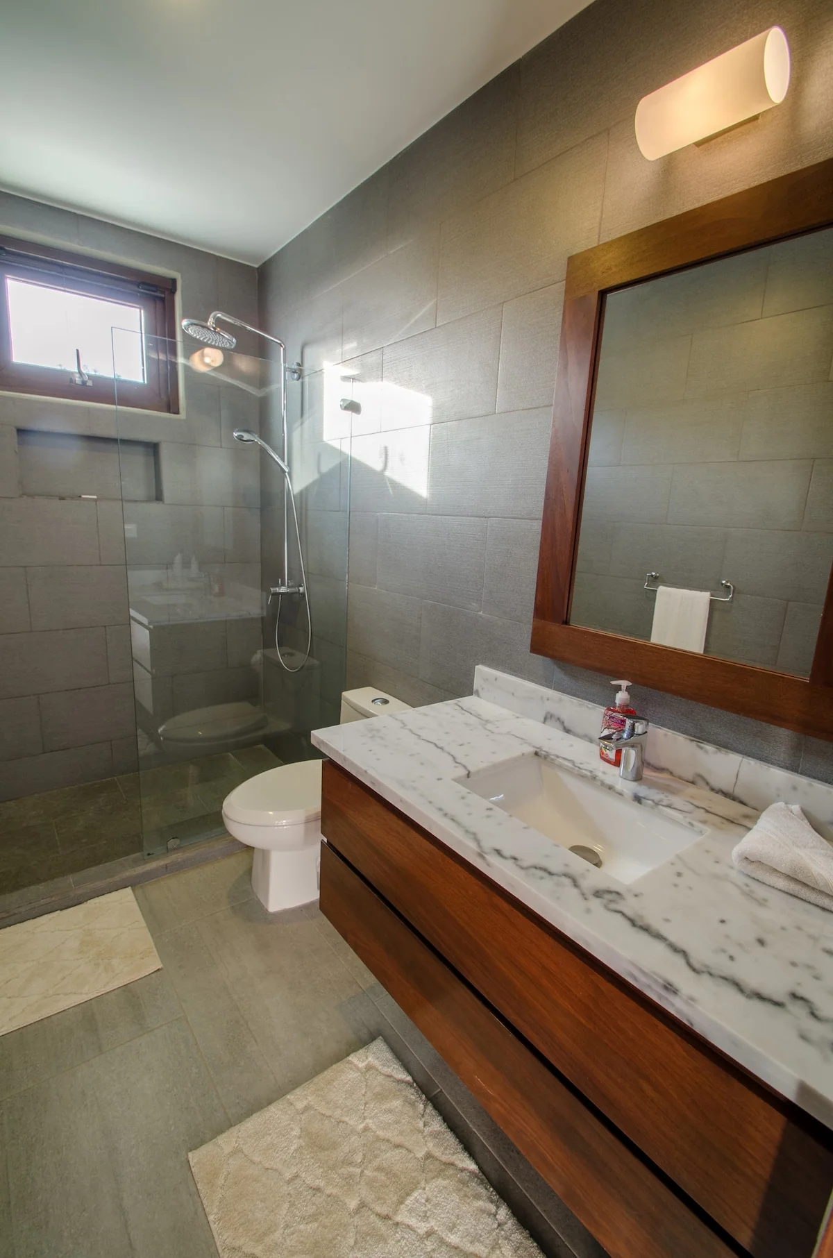 A modern bathroom with a glass-enclosed shower, toilet, large wood-framed mirror, and marble vanity illuminated by daylight.