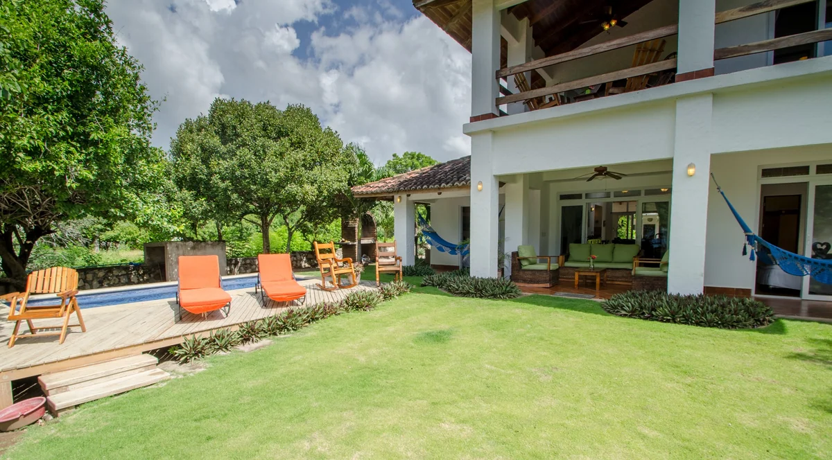 A bright garden lawn beside a white house with patio chairs, hammocks, trees, and a stone-edged terrace.