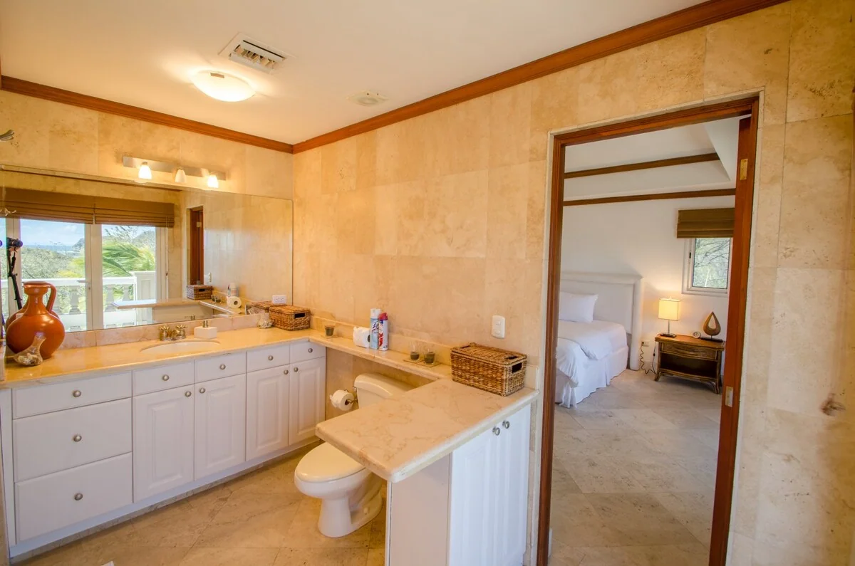 A spacious bathroom with marble-look walls, white cabinetry, a bathtub, and an open doorway to a bedroom.