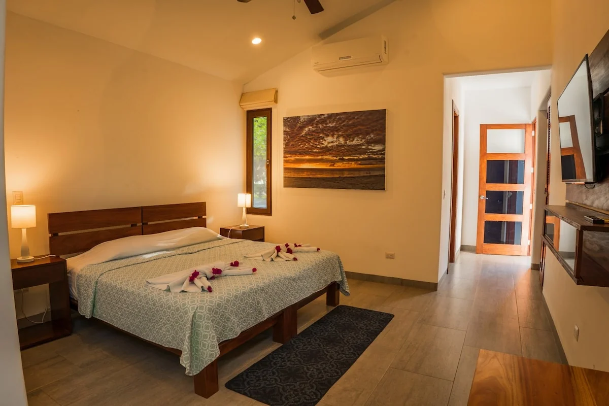 A warmly lit bedroom contains a large bed, bedside lamps, wall art, an air conditioner, and an open doorway.