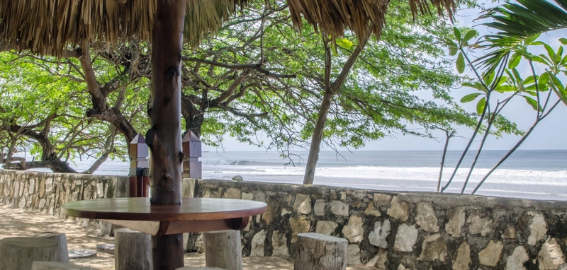 The image depicts a cozy seating area under a thatched roof near a beach. The seating area consists of wooden tables and stools. In the background, there is a stone wall, beyond wh