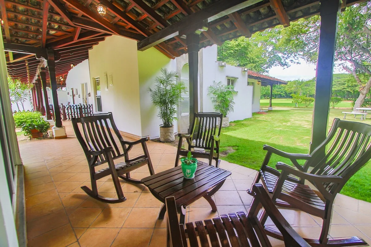 A covered tiled terrace furnished with wooden rocking chairs and a small table overlooks a green lawn and garden.
