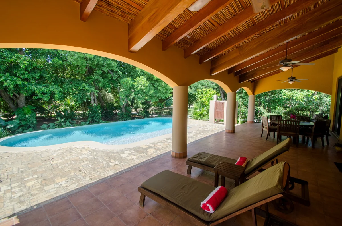 A covered outdoor terrace with wooden beams, lounge chairs, a dining table, and a swimming pool beside lush greenery.