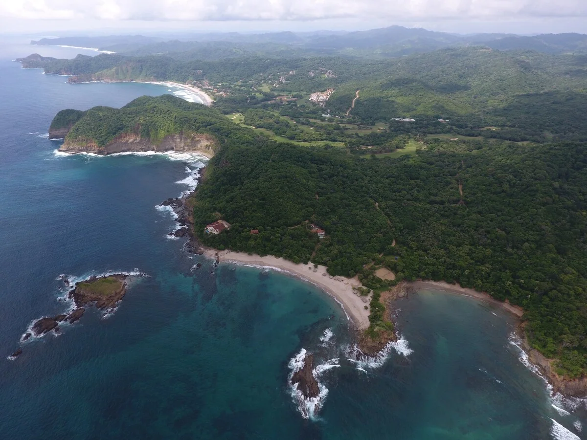 An aerial view shows a forested coastline with turquoise water, rocky cliffs, sandy coves, small offshore rocks, and scattered buildings.