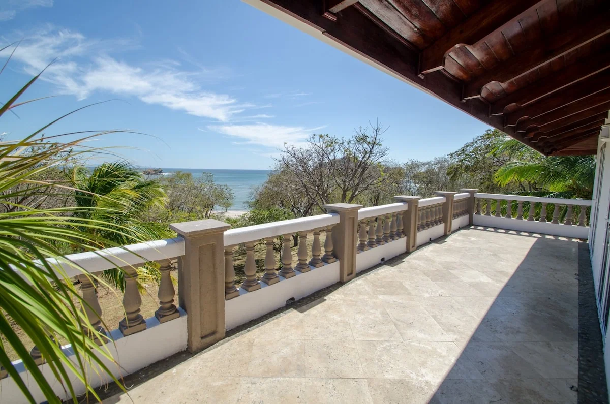 A sunny tiled terrace with a balustrade overlooks trees and the ocean.
