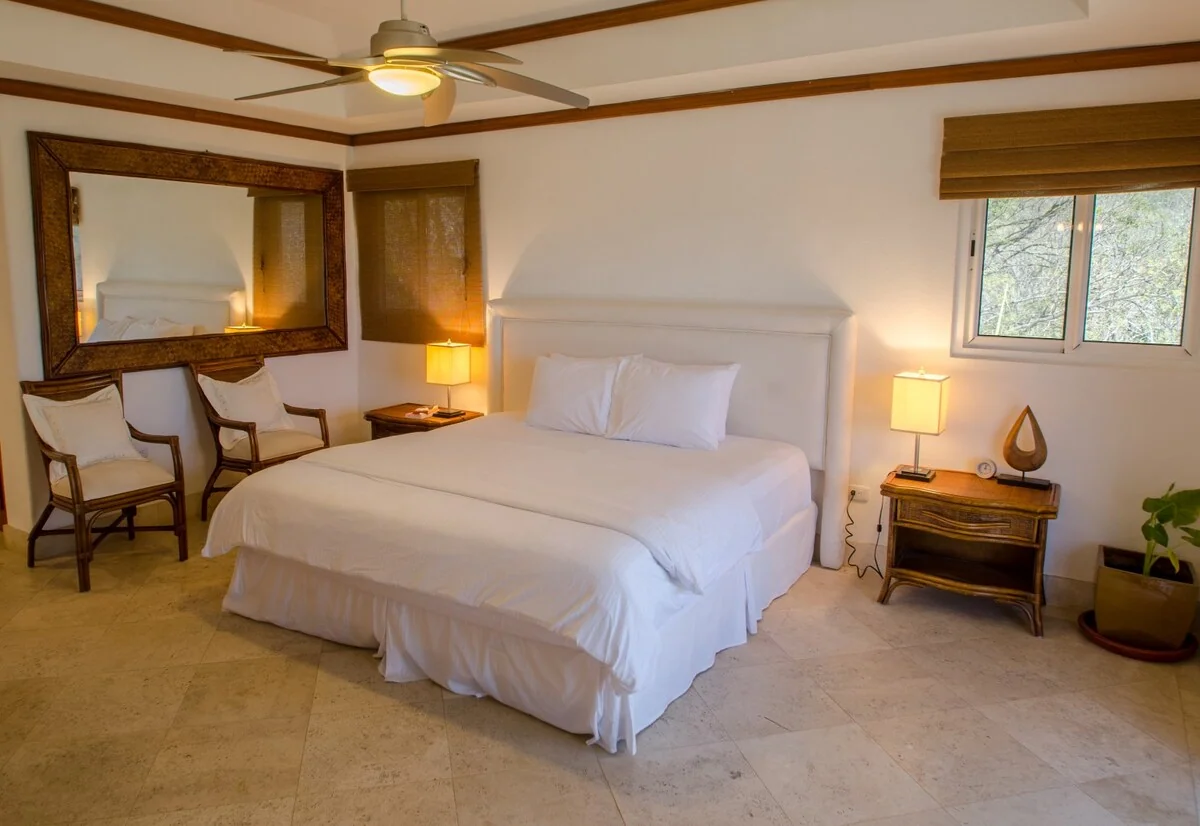 A bright bedroom contains a large white bed, two chairs, bedside lamps, a wall mirror, and a ceiling fan.