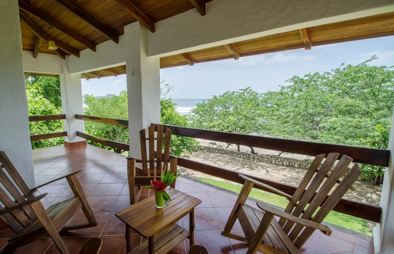 The image shows a covered veranda with a wooden ceiling, featuring several wooden chairs and a small wooden table with a vase of red flowers. In the background, lush green trees an