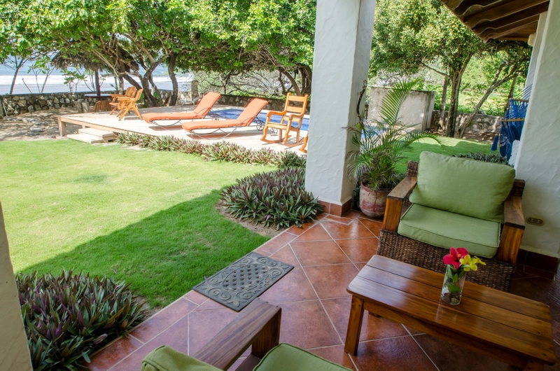 The image shows an outdoor patio area furnished with green cushioned chairs and a wooden table. Beyond the patio, there is a lush grassy area bordered by decorative plants. In the 