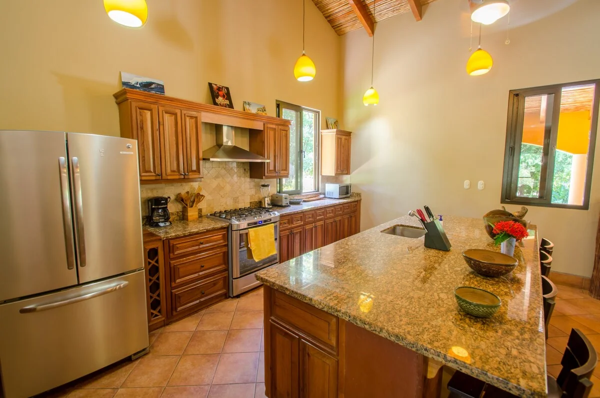A spacious kitchen with wooden cabinetry, stainless steel appliances, pendant lights, and a large granite island.