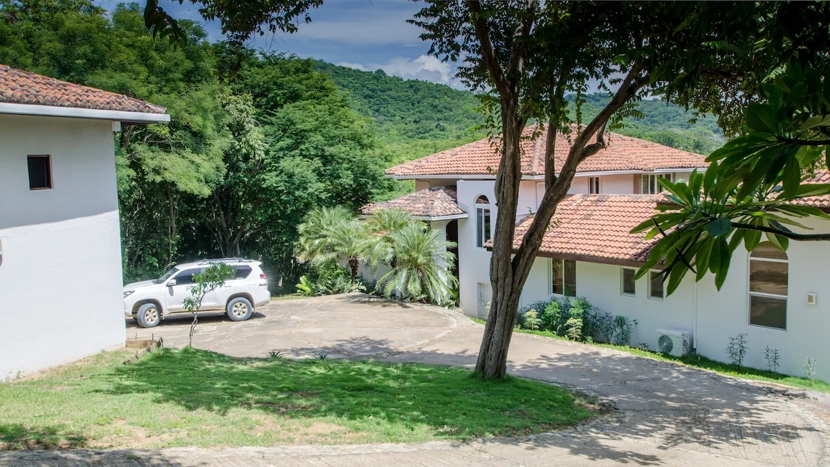 A tiled-roof house and driveway sit among trees and palms with green hills visible behind.