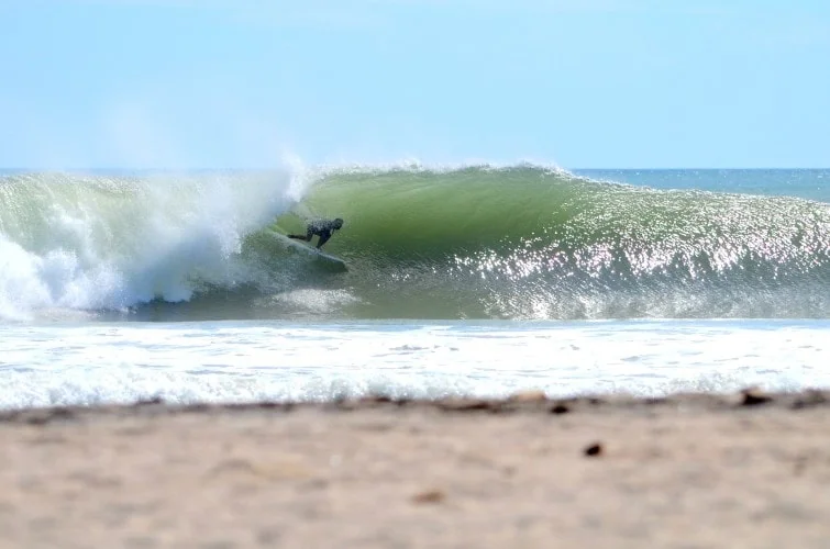A surfer rides inside a breaking green wave near a sandy beach under a clear blue sky.