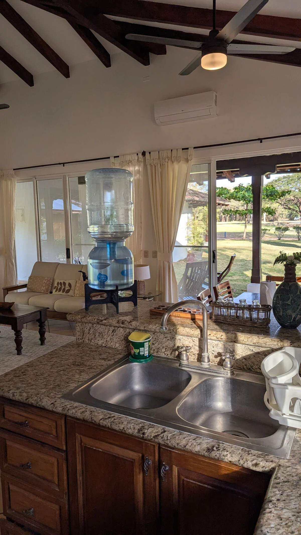 A kitchen with a granite island, double sink, water cooler, wooden cabinets, and large windows overlooking a lawn.