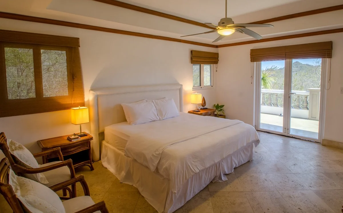 A warmly lit bedroom with a large neatly made bed, wooden furnishings, lamps, and glass doors opening to a balcony.