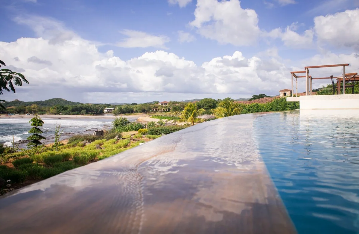 An infinity pool overlooks a green shoreline and distant houses beneath a bright, cloud-filled sky.