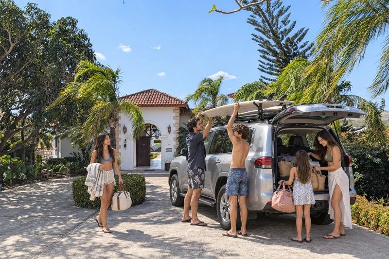 Six people gather beside an SUV with surfboards and luggage in front of a bright house surrounded by palm trees.