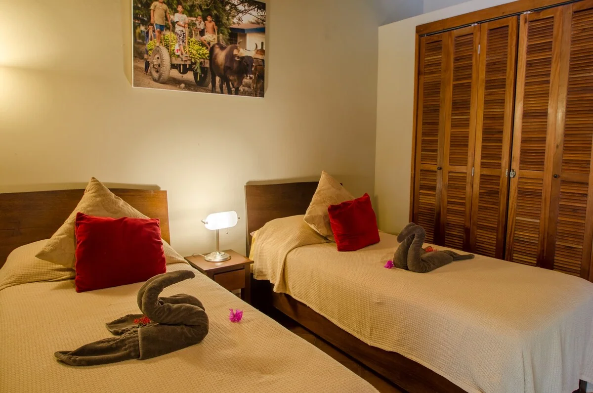 A warmly lit bedroom contains two single beds with beige linens, red accent pillows, folded towel decorations, a bedside lamp, and a wooden louvered wardrobe.