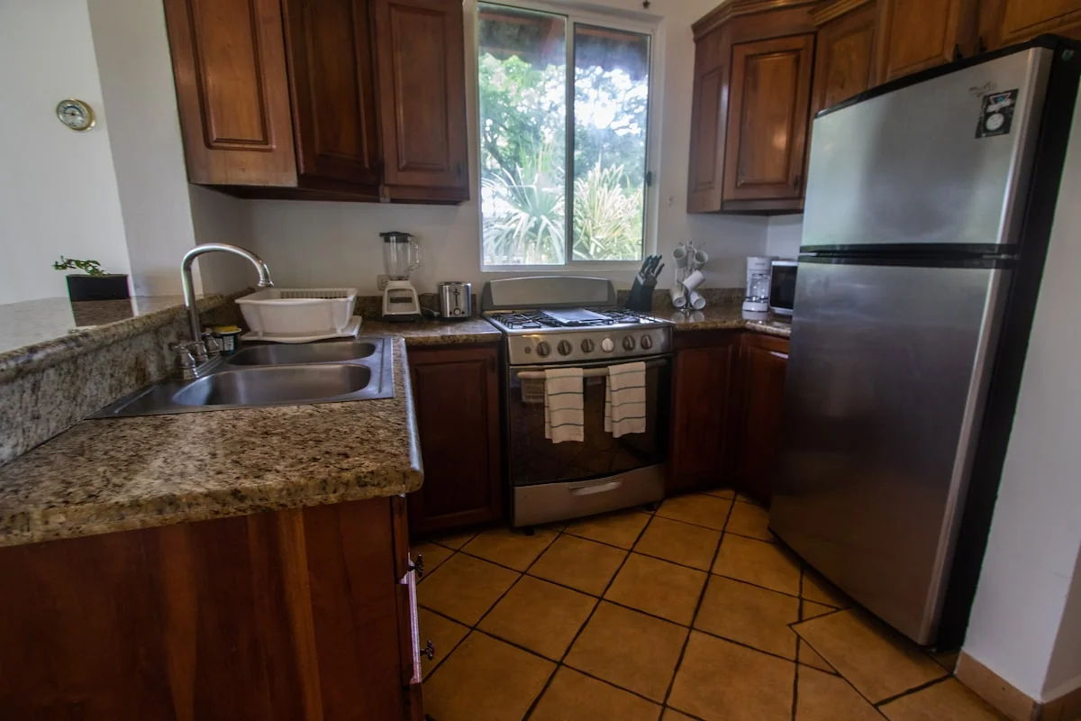 A warm-toned kitchen with wooden cabinets, granite countertops, a sink, stove, refrigerator, and a window overlooking greenery.