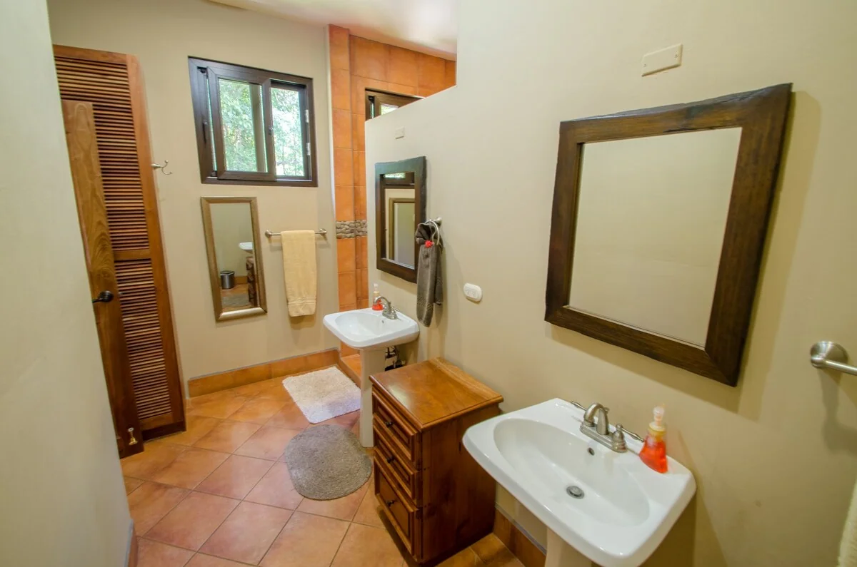 A warm-toned bathroom with two sinks, large mirrors, wooden cabinetry, towels, and a window.
