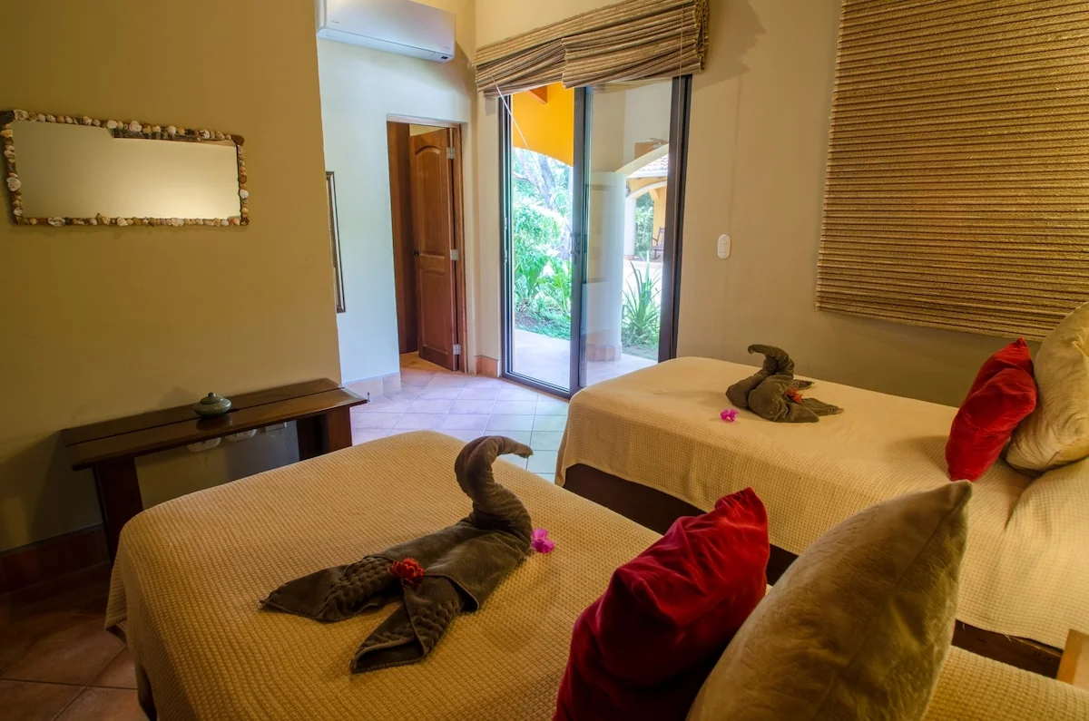 A warmly lit bedroom contains two neatly made beds with folded towel decorations, red cushions, wooden furniture, and an open glass door to greenery.