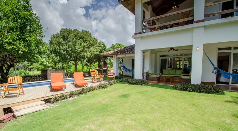 The image features a cozy outdoor scene with a well-maintained lawn and a poolside deck. On the deck, there are several wooden chairs and two orange lounge chairs. The house in the