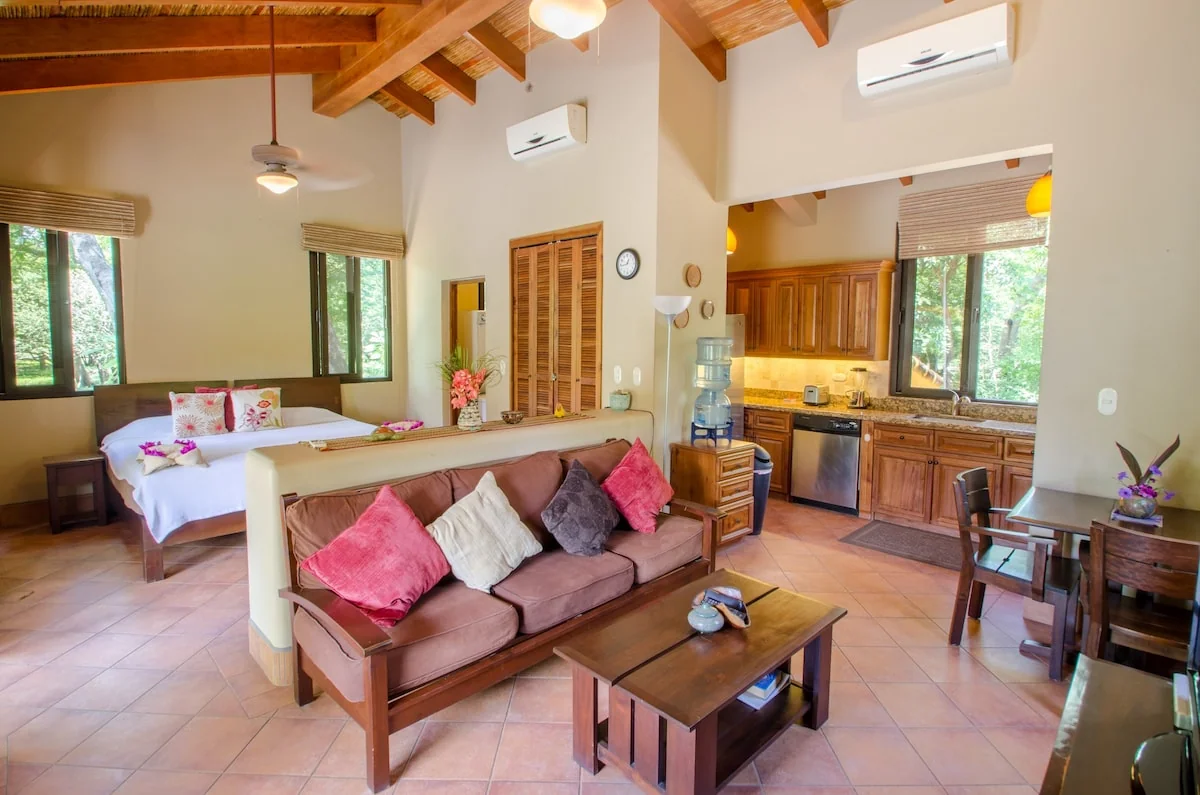A bright open-plan interior contains a sofa, coffee table, bed, and wood-paneled kitchen beneath exposed wooden beams.