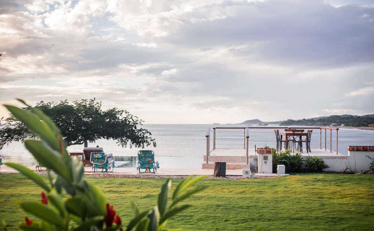 A grassy waterfront area with lounge chairs, a railing, plants, and a calm sea beneath a cloudy sky.