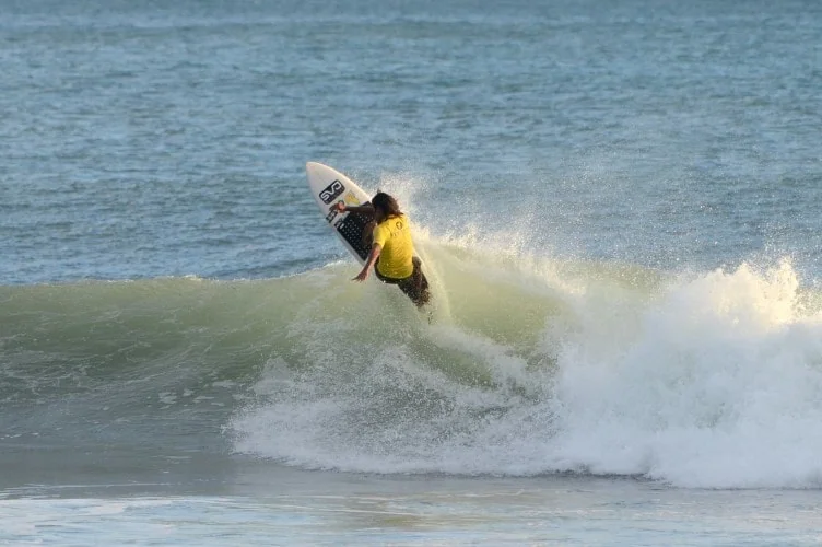A surfer in a yellow shirt rides a breaking ocean wave while holding a surfboard.