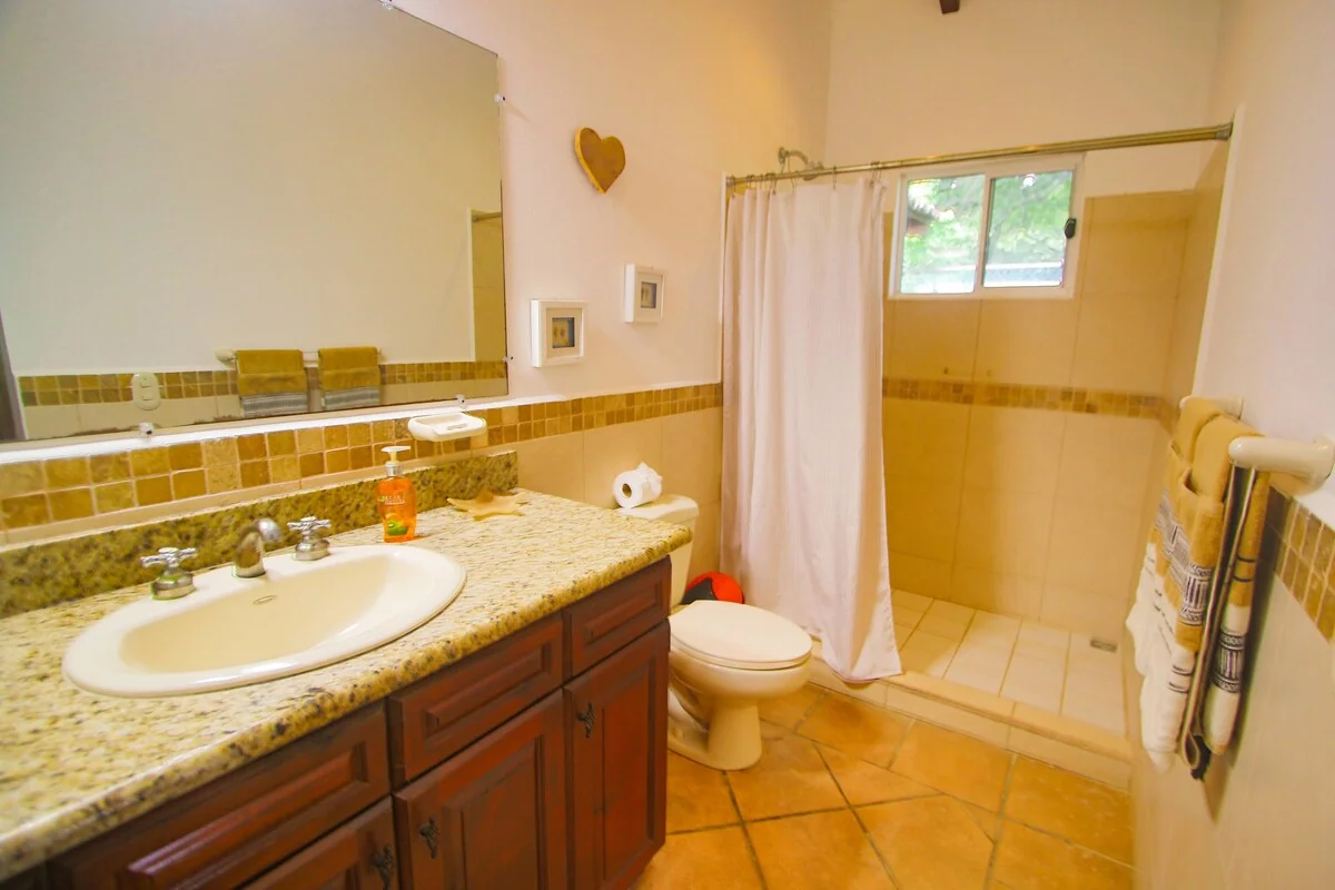 A bright bathroom with a granite vanity, large mirror, toilet, tiled shower, and hanging towels.