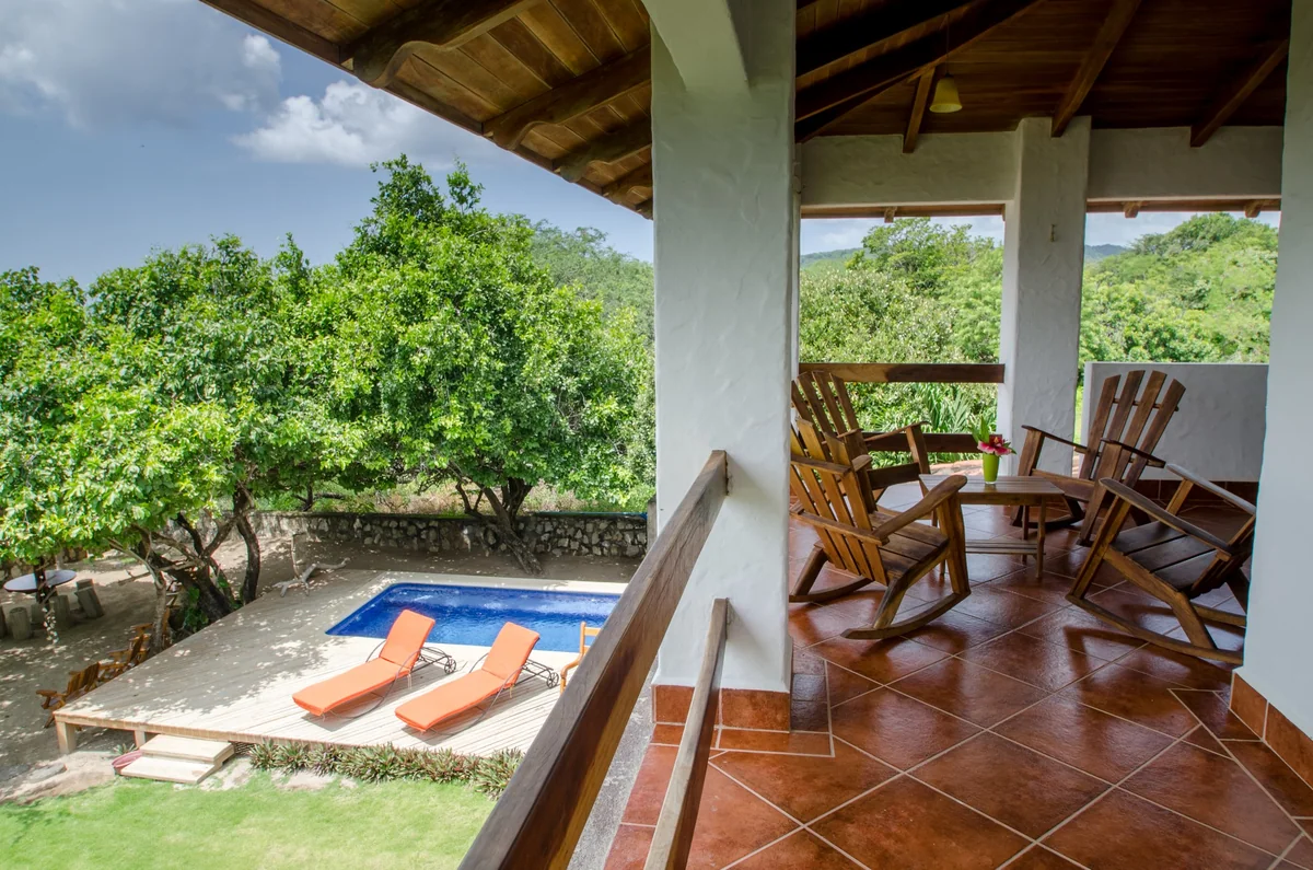 A covered terrace with wooden rocking chairs overlooks a small blue pool, lounge chairs, and leafy trees.