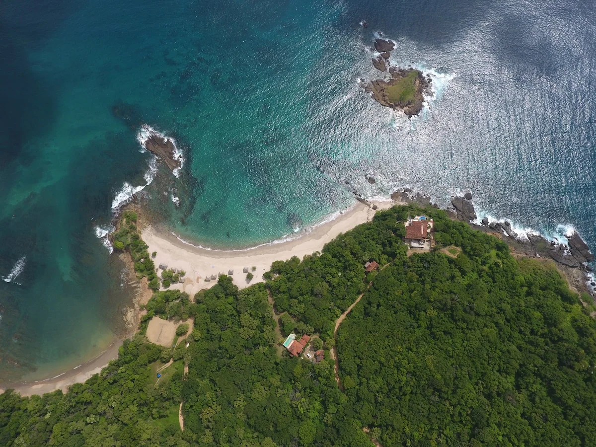 An aerial view shows a curved sandy beach, turquoise ocean, rocky islets, and dense green coastal forest with a few buildings.