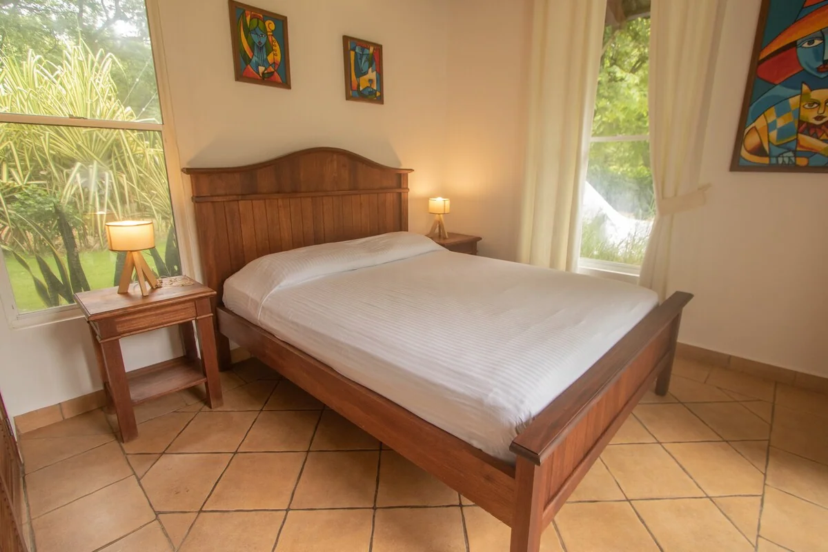 A wooden bed with white bedding stands in a tiled bedroom with bedside lamps, windows, and colorful wall artwork.