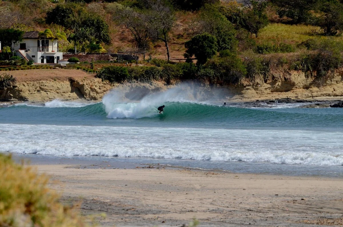 Two surfers are visible in breaking waves near a sandy beach with a house and vegetation in the background.