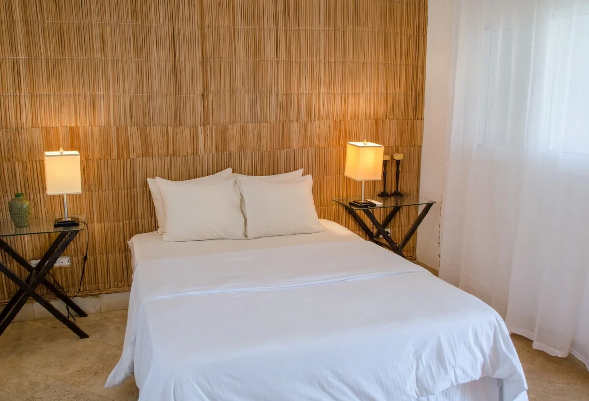 A neatly made bed with white linens sits against a warm woven wall, flanked by lit table lamps and dark folding tables.