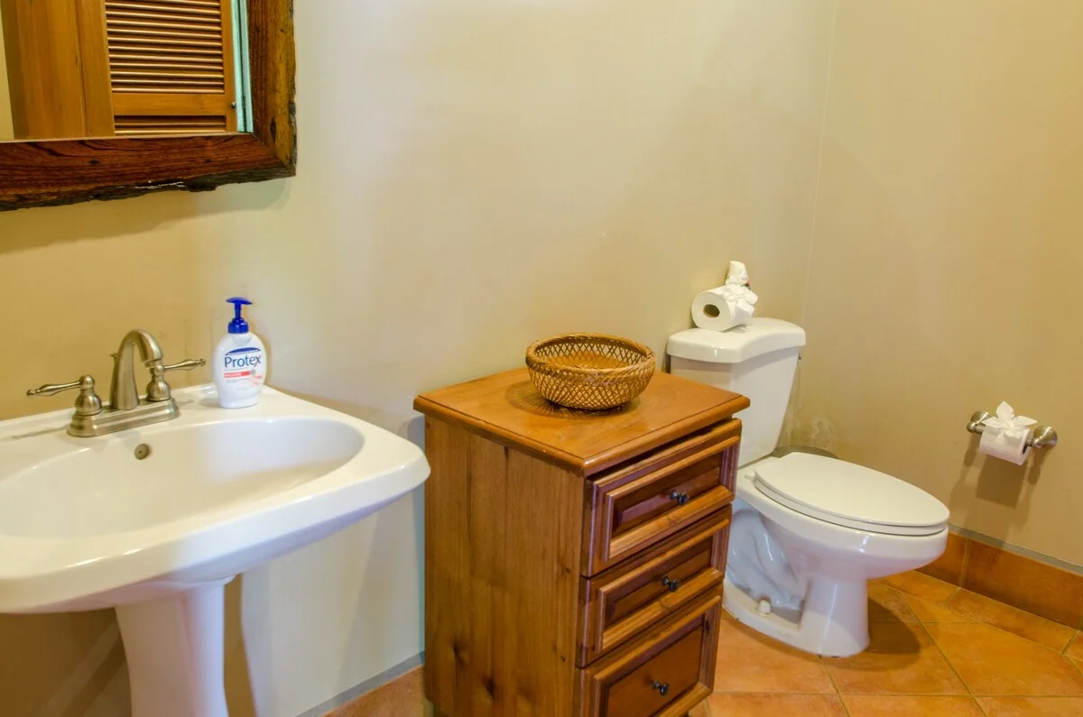 A bright bathroom contains a pedestal sink, toilet, wooden cabinet, woven basket, and shuttered window.
