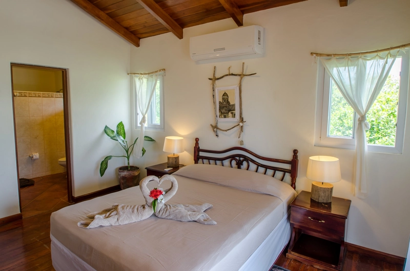 A cozy bedroom featuring a wooden ceiling and furnished with a comfortable bed adorned with creatively folded towels in the shape of swans. Warm natural light pours in through two 
