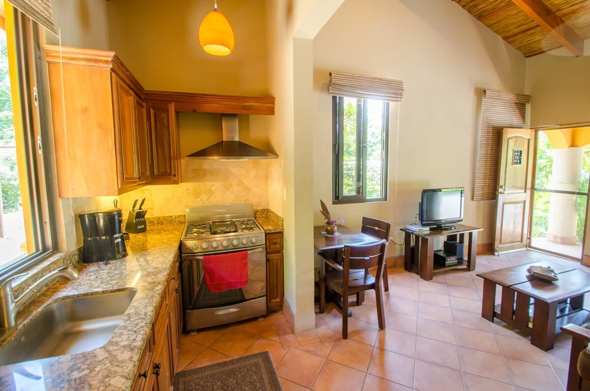 A warmly lit open-plan kitchen and living area with wooden cabinetry, a stove, dining table, television, and seating.