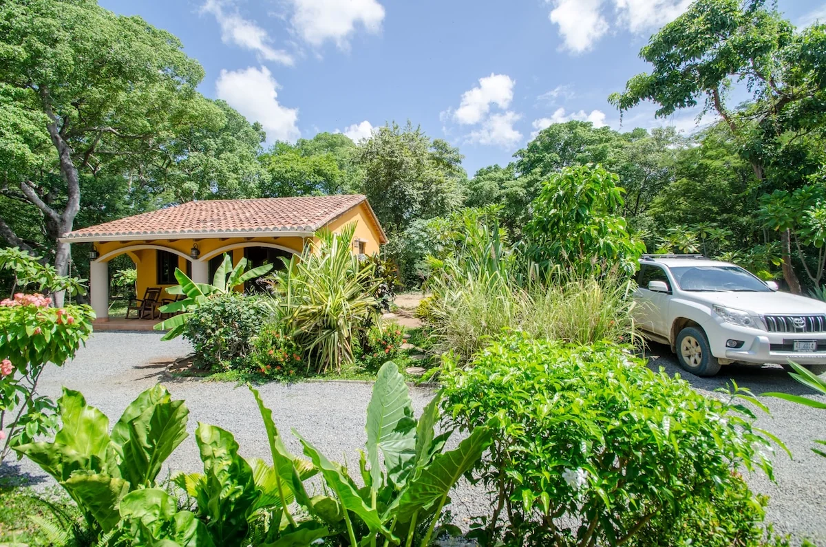 A small tiled-roof house is surrounded by lush tropical plants and trees, with a white SUV parked on the driveway.