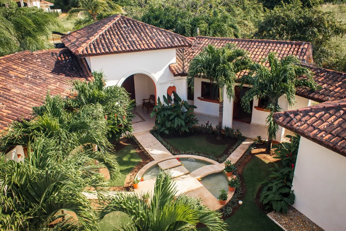 An elevated view shows a white stucco courtyard house with terracotta tiled roofs, lush palms, paved walkways, and a small central pond.