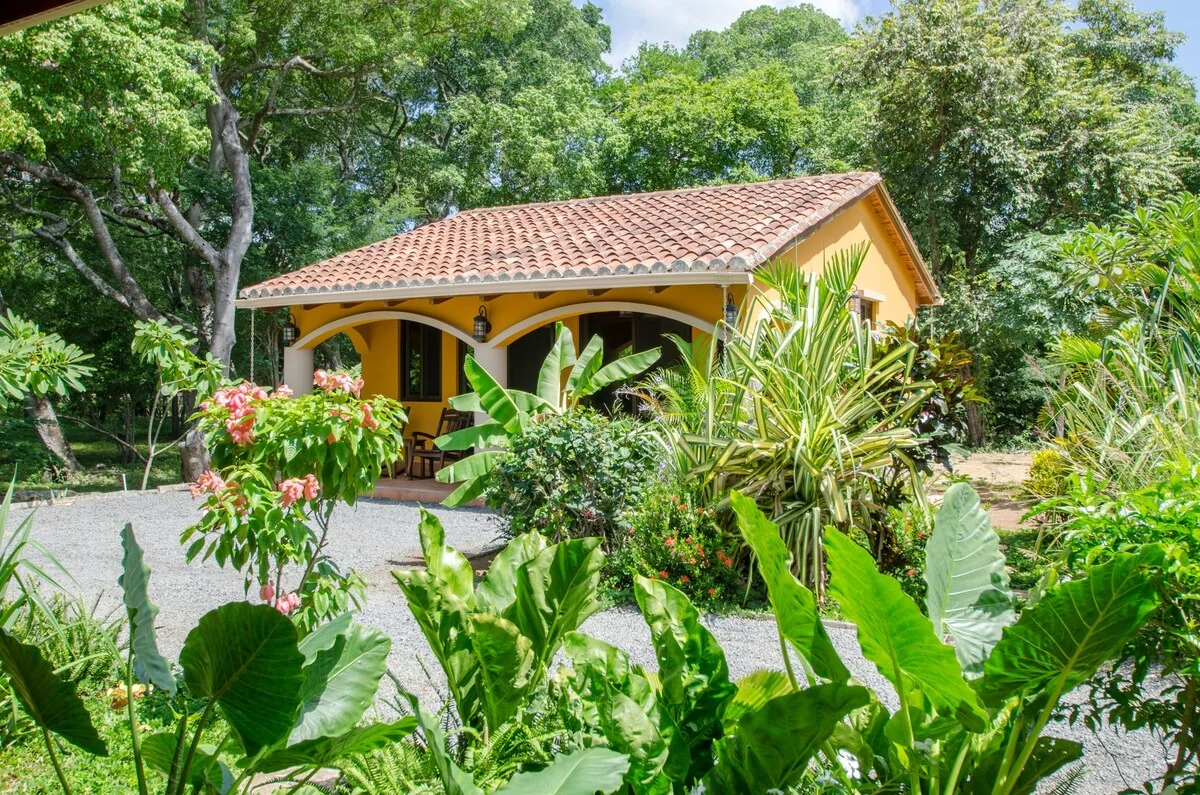 A tiled-roof bungalow with an open covered porch is surrounded by lush tropical plants and trees.